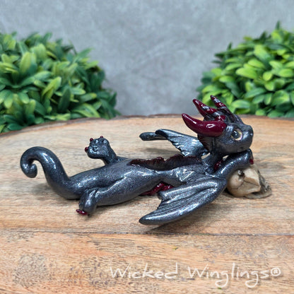 A clay dragon with a skull against a wood surface with greenery. Includes text Wicked Winglings