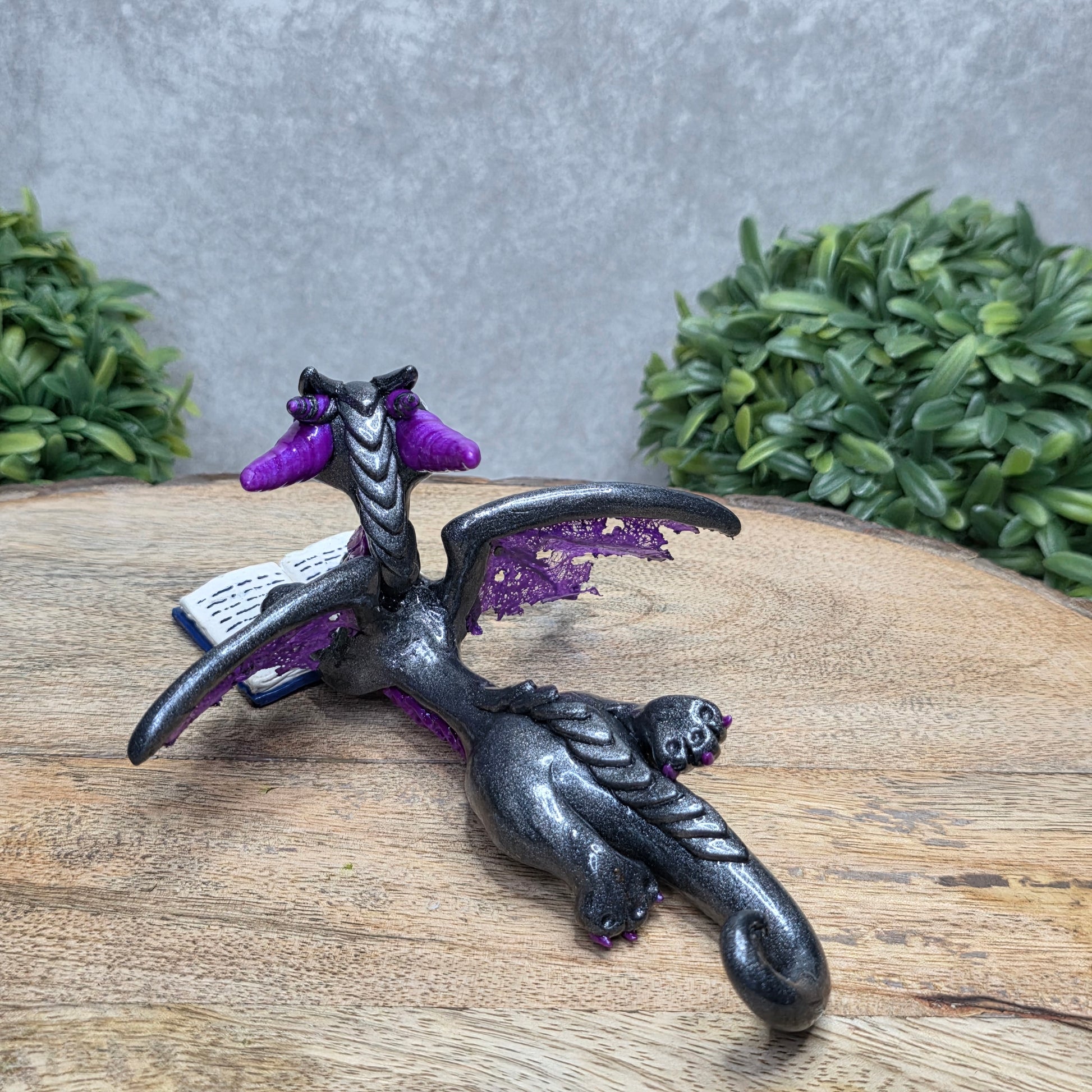 A dragon reading a book against a wood surface with greenery
