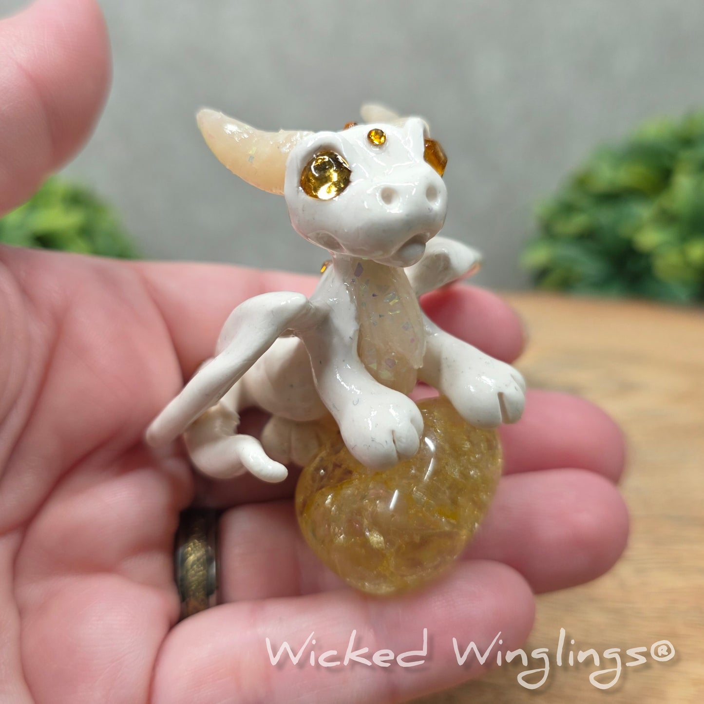 A hand holding a baby clay dragon sculpture with a crackled quartz against a wood background with greenery and text Wicked Winglings