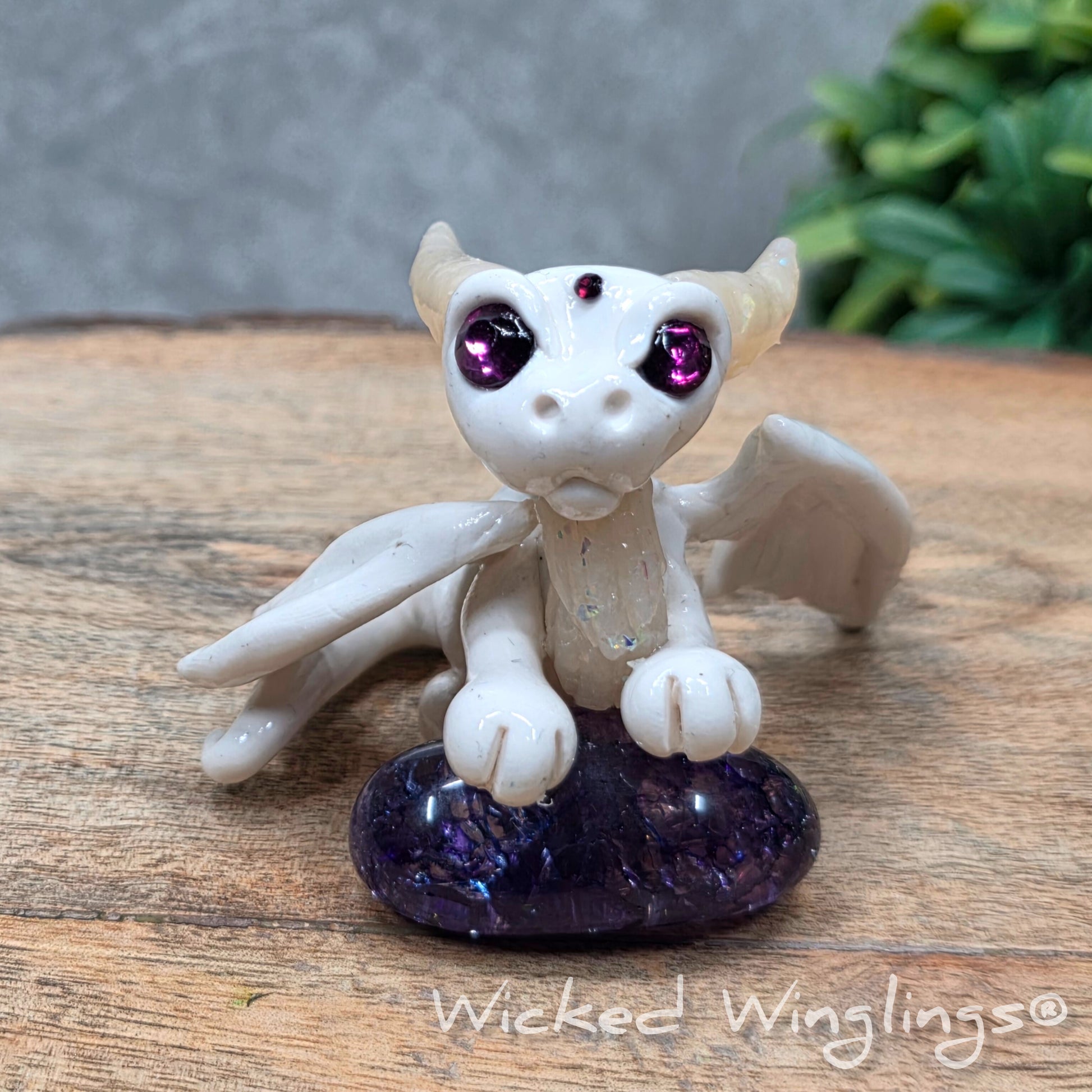 A baby clay dragon sculpture with a crackled quartz against a wood background with greenery and text Wicked Winglings