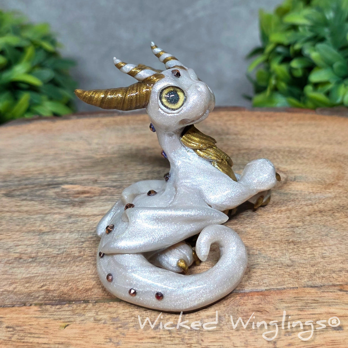  A white clay dragon sitting against a blurred background with greenery. Text says Wicked Winglinng.