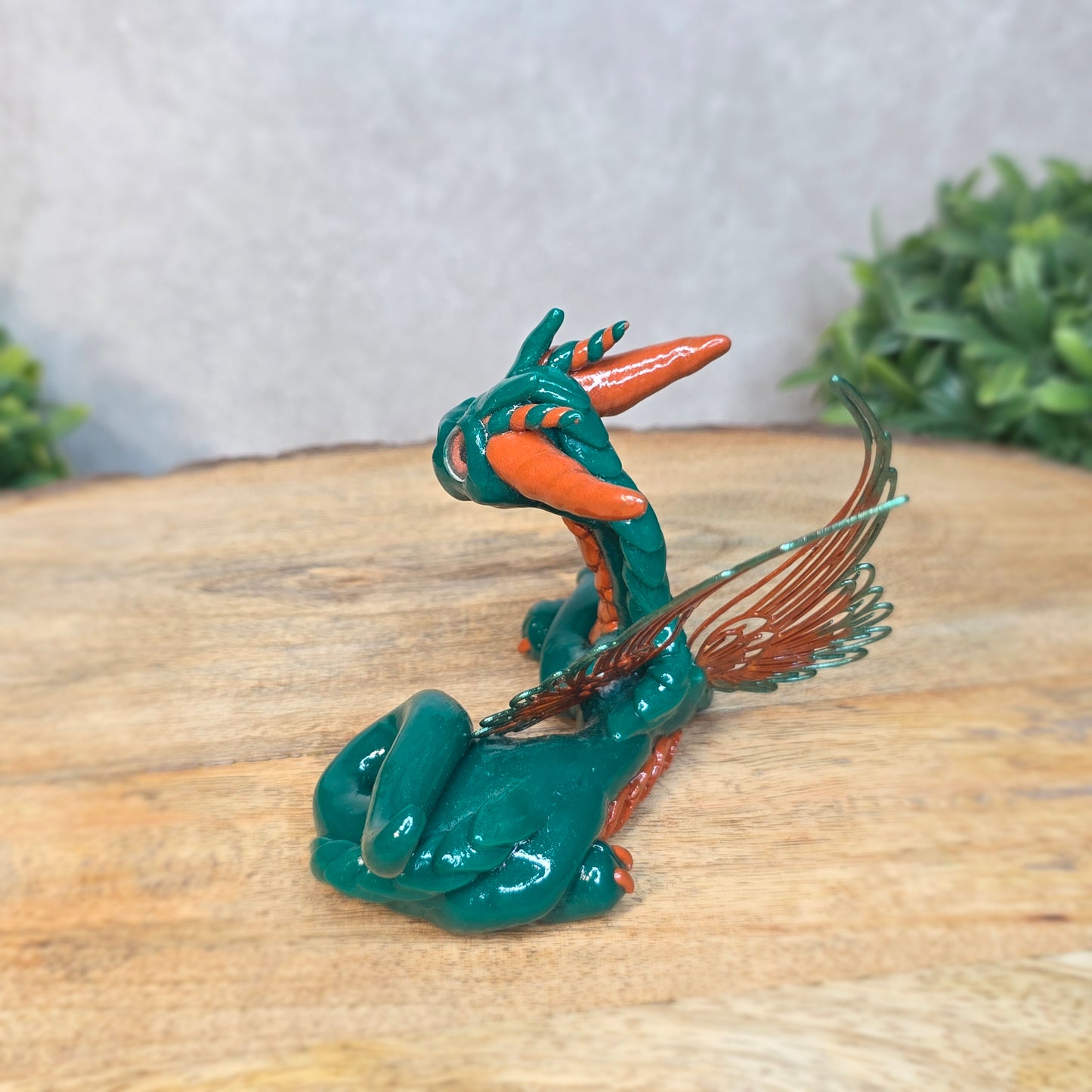 Green and orange dragon figurine on a wooden surface with a blurred green plant in the background