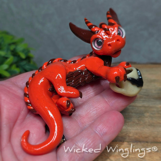 A hand holding an orange clay dragon with a bat tea cup against a wood surface with greenery. Text reads Wicked Winglings.