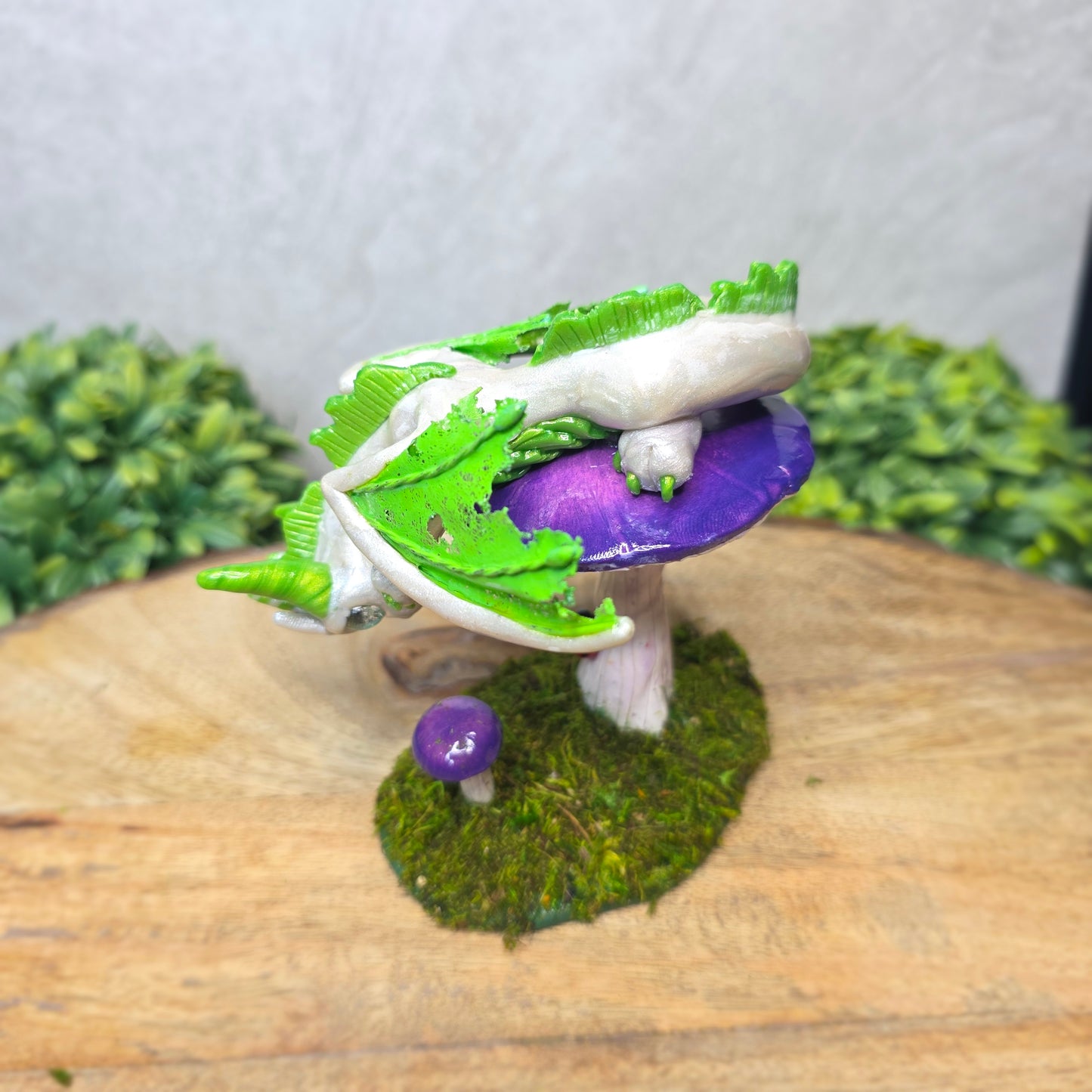 A clay dragon figurine on a  mushroom on a wooden surface with greenery.