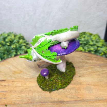 A clay dragon figurine on a  mushroom on a wooden surface with greenery.