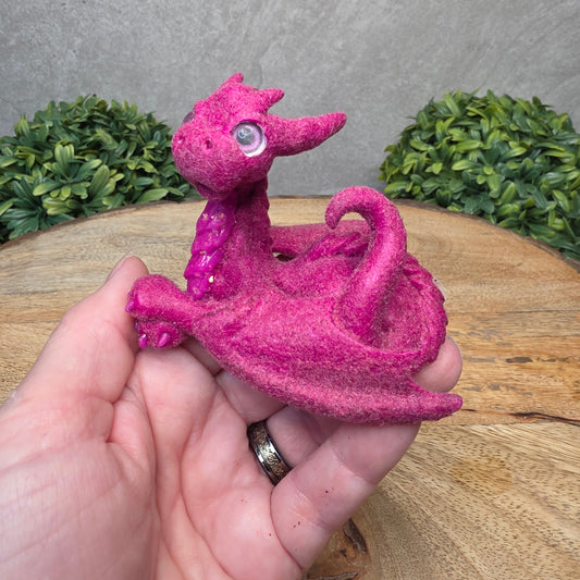 A hand holding a flocked clay dragon sculpture on a wood surface with greenery.