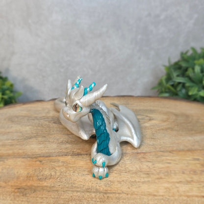 A white and teal clay dragon against a blurred wood surface