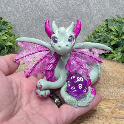 Hand holding a clay dragon with a D20 dice against a wood surface with greenery.