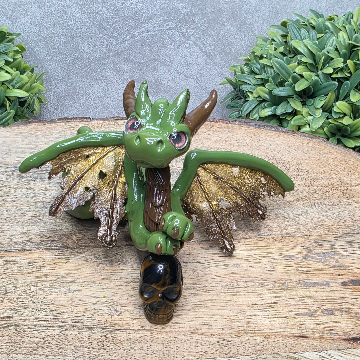 A clay dragon with a tigers eye crystal skull against a wood surface with greenery.