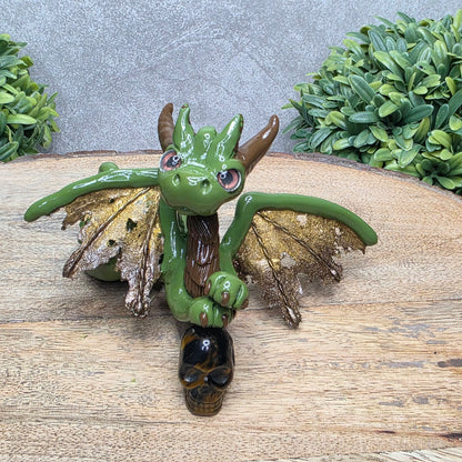 A clay dragon with a tigers eye crystal skull against a wood surface with greenery.