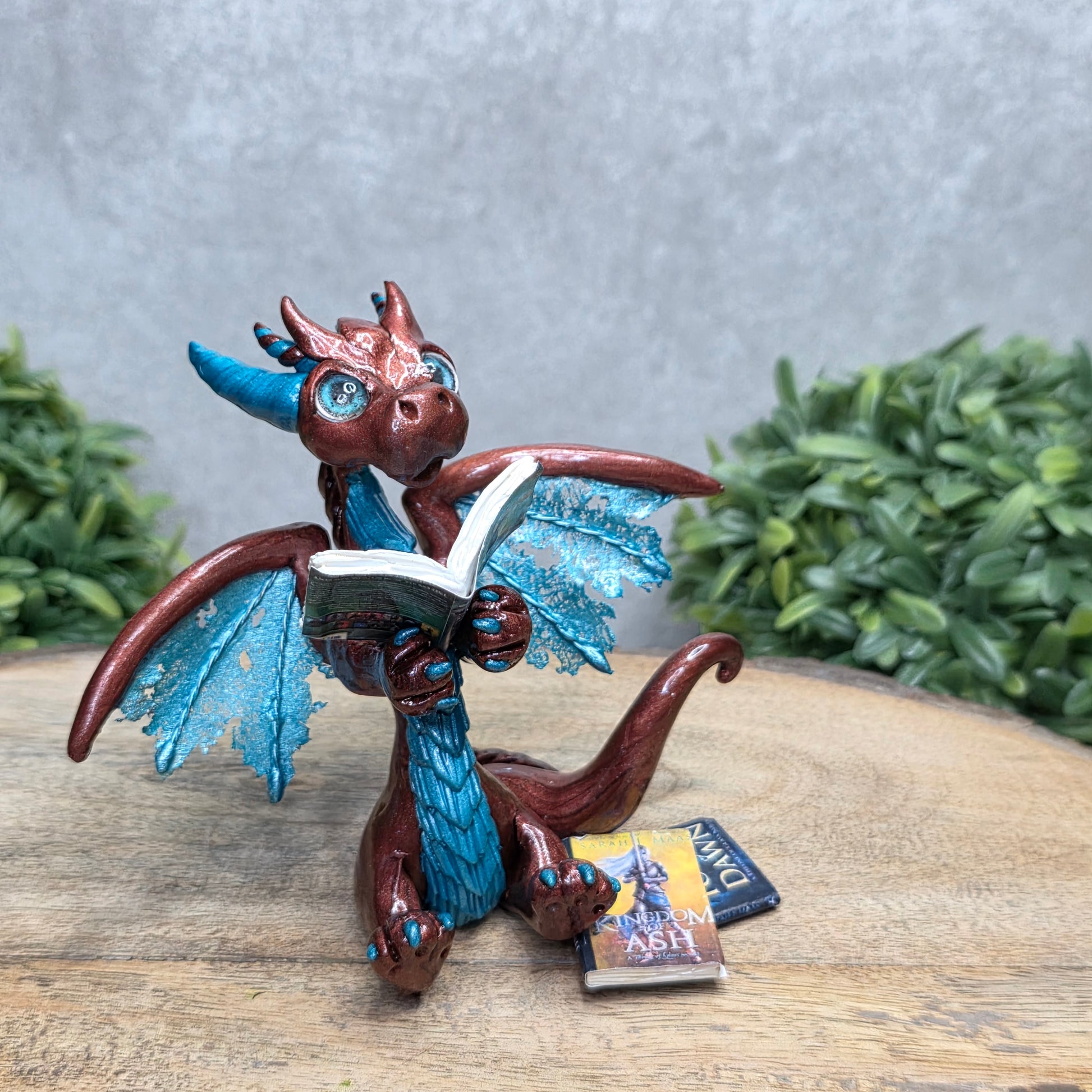 A dragon reading a book against a wood surface with greenery