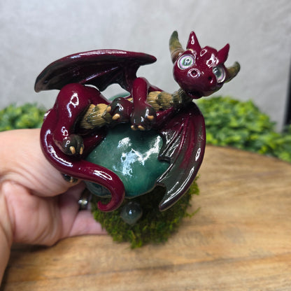 Clay dragon figurine held by a hand with a wooden surface and greenery in the background