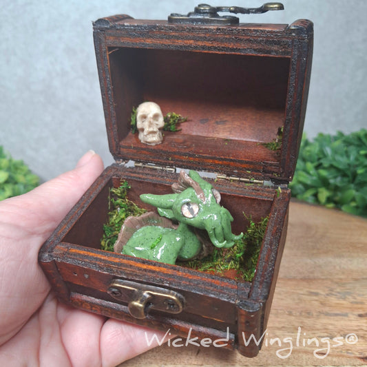 A hand holding a clay dragon in a chest accented with moss and a skull. Text says Wicked Winglings