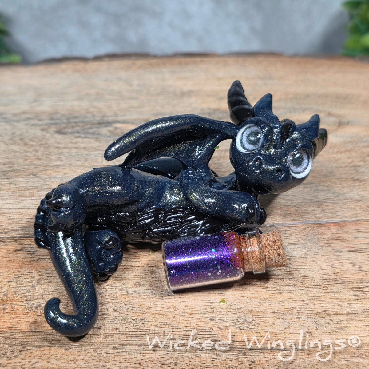 A clay dragon with a potion bottle on a wood surface. Text reads Wicked Winglings.