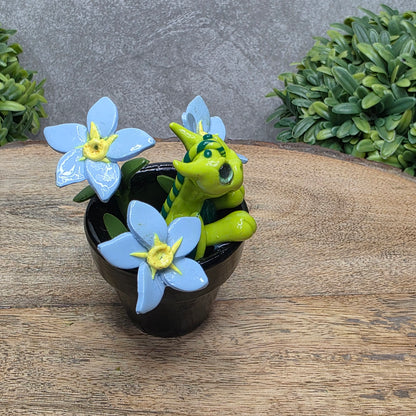 Side view of a clay baby dragon in a pot with forget me nots against a wood surface with greenery.