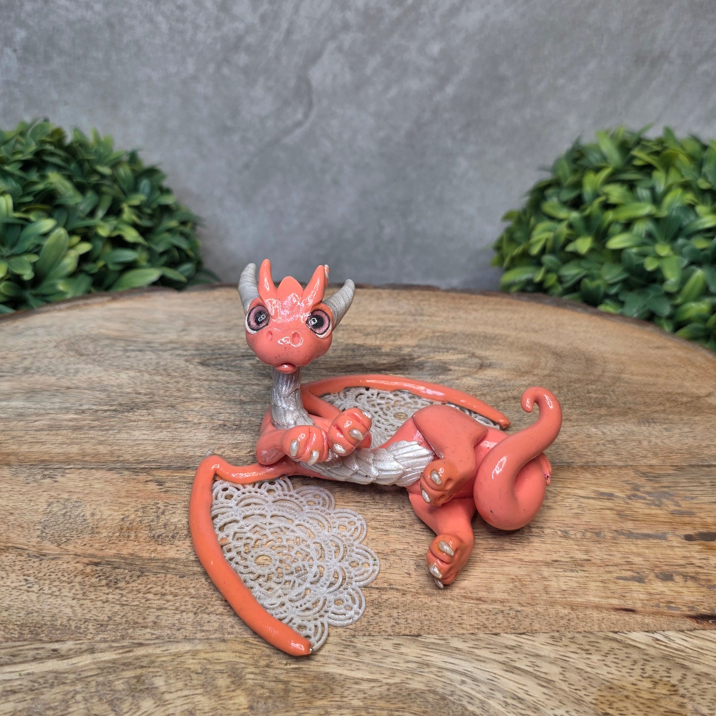 A clay dragon sculpture with lacy wings on a wood surface with greenery.