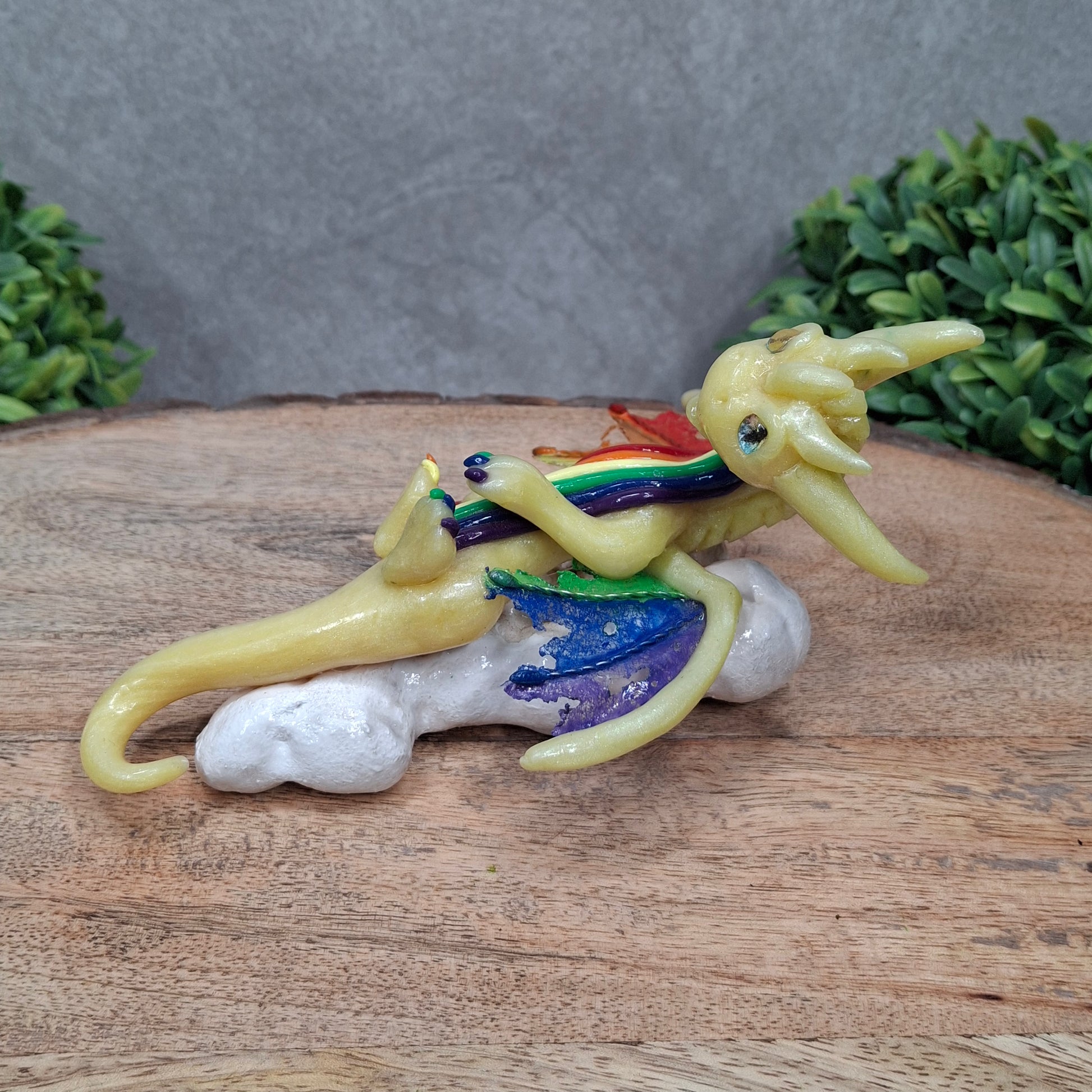 A yellow dragon with rainbow accents on a cloud sitting against a wood surface with greenery