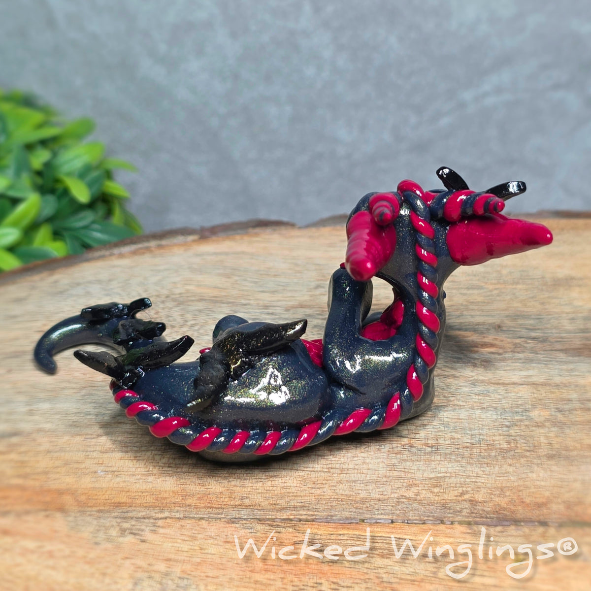 Back view of a clay dragon with bats sitting on a wood surface with greenery. Text reads Wicked Winglings.