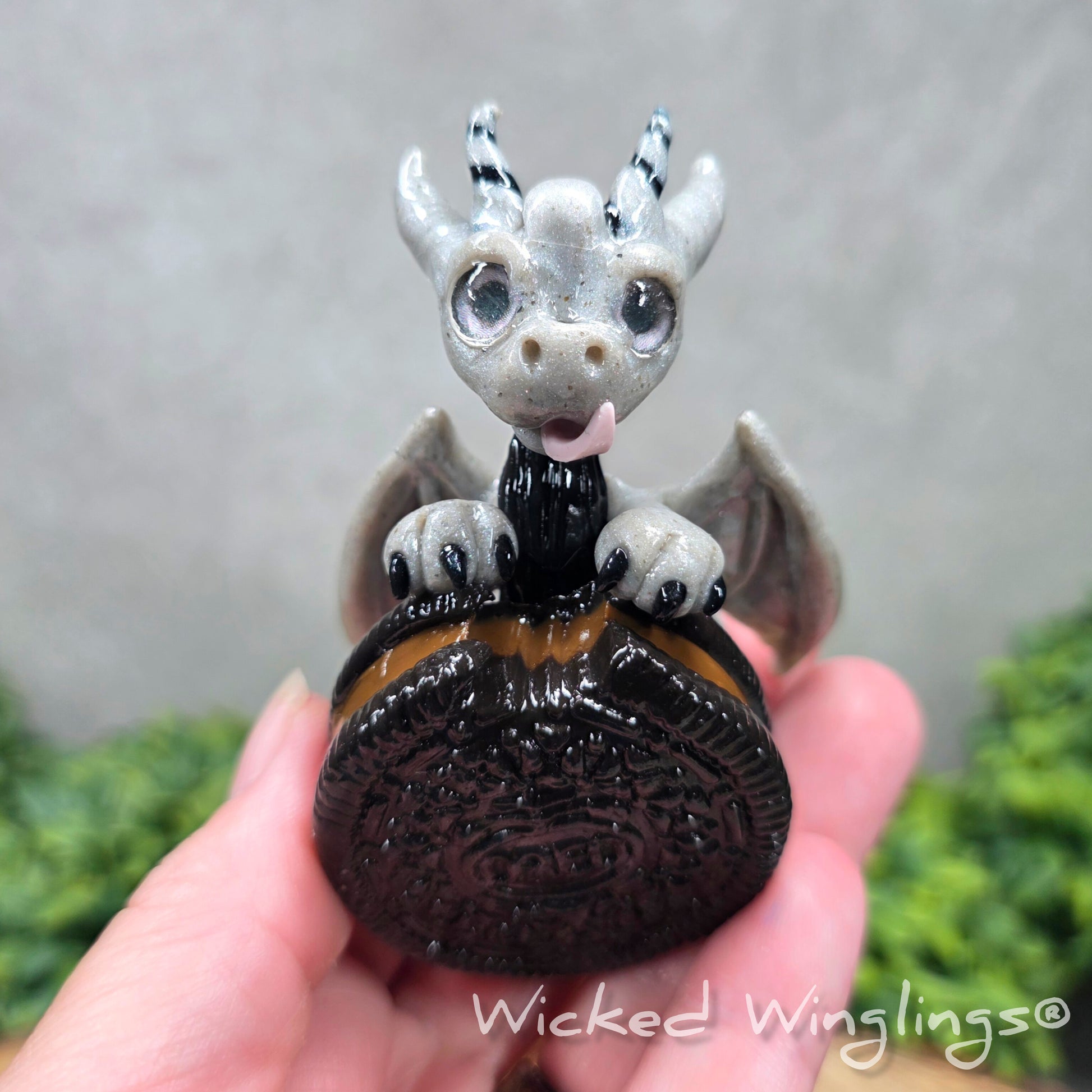 A hand holding a clay dragon with a cookie with a Wicked Winglings® watermark.