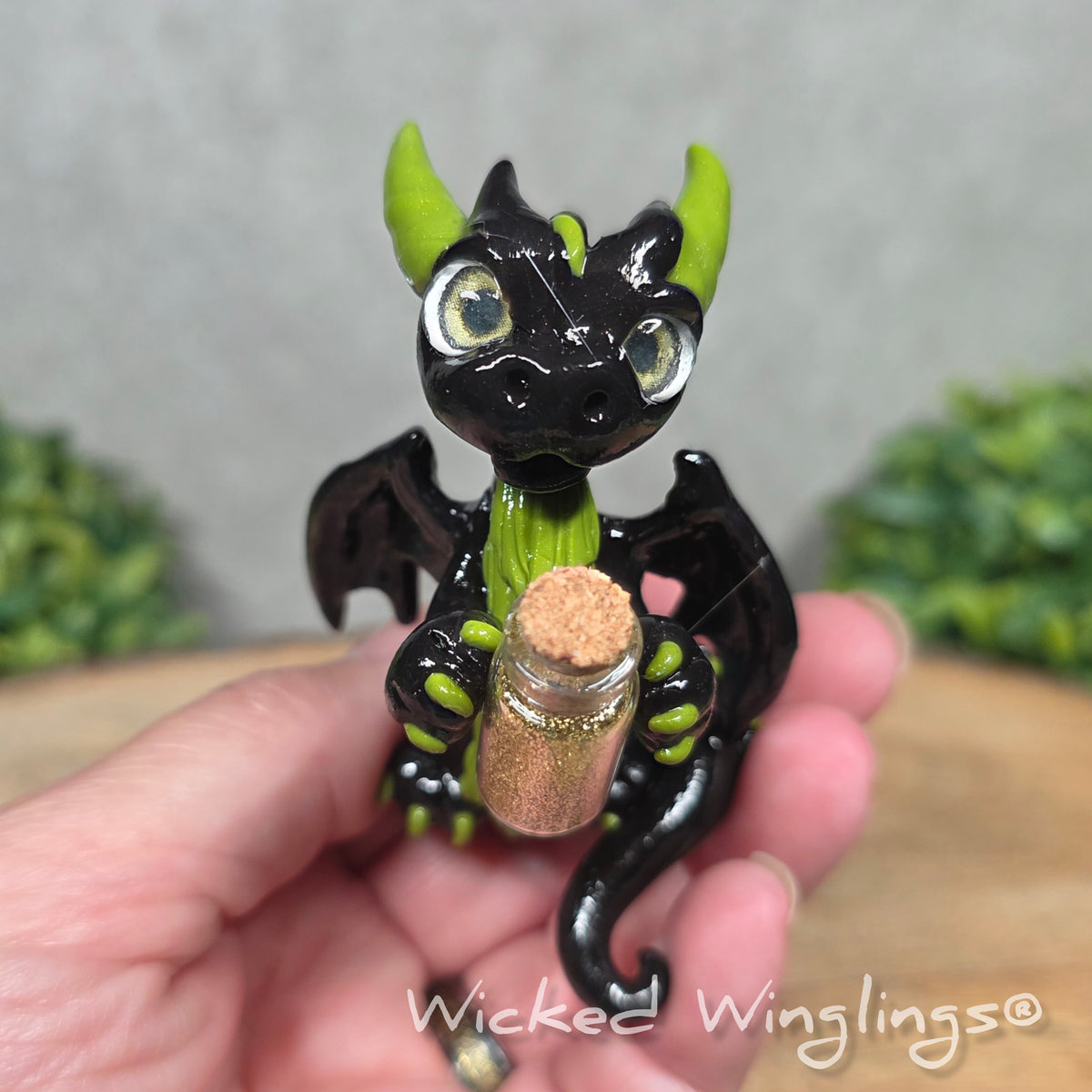 A hand holding a clay dragon with a potion bottle on a wood surface. Text reads Wicked Winglings.