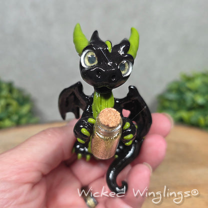 A hand holding a clay dragon with a potion bottle on a wood surface. Text reads Wicked Winglings.