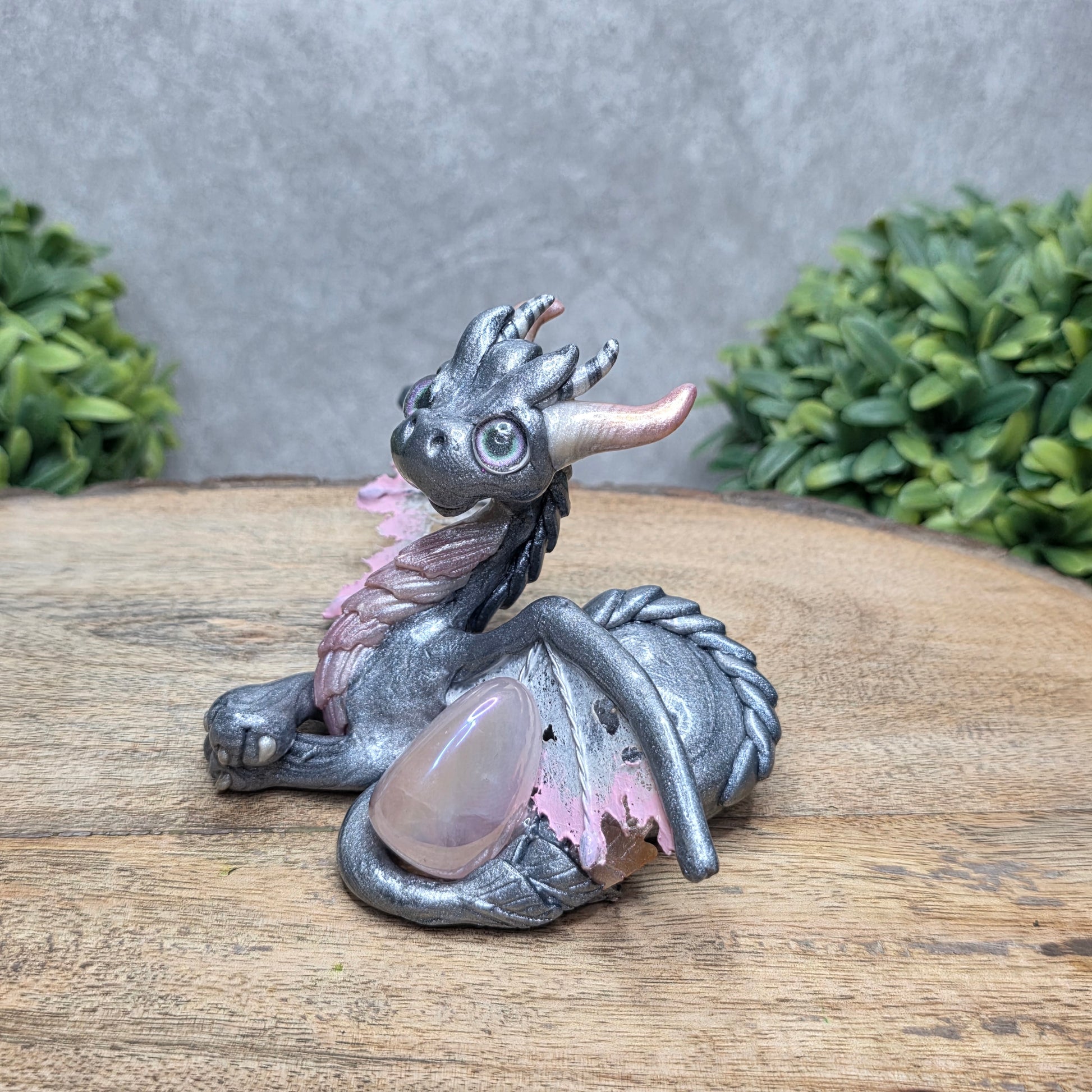 Side view of a clay dragon with a rose quartz crystal against a wood surface with greenery.