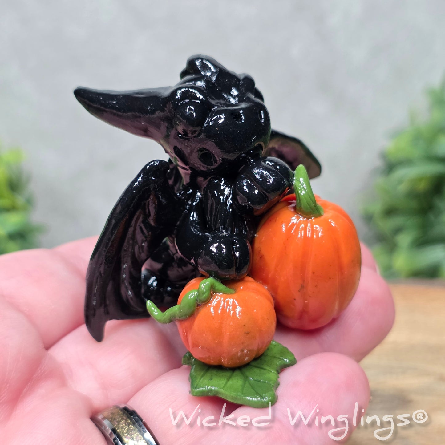 A hand holding a baby clay dragon sculpture with pumpkins against a wood background with greenery and text Wicked Winglings