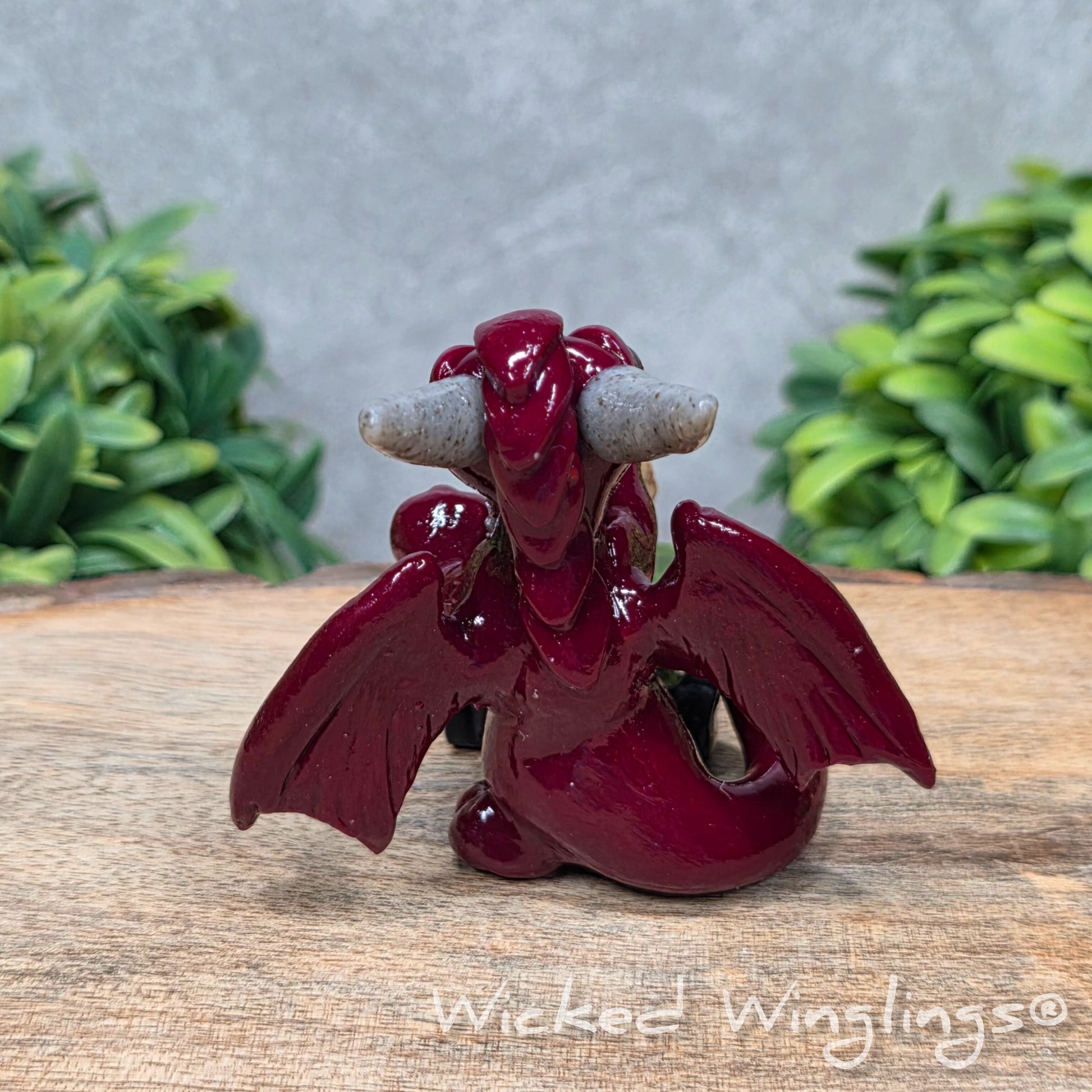 A clay dragon with a skull and books on a wood surface with greenery. Includes text Wicked Winglings