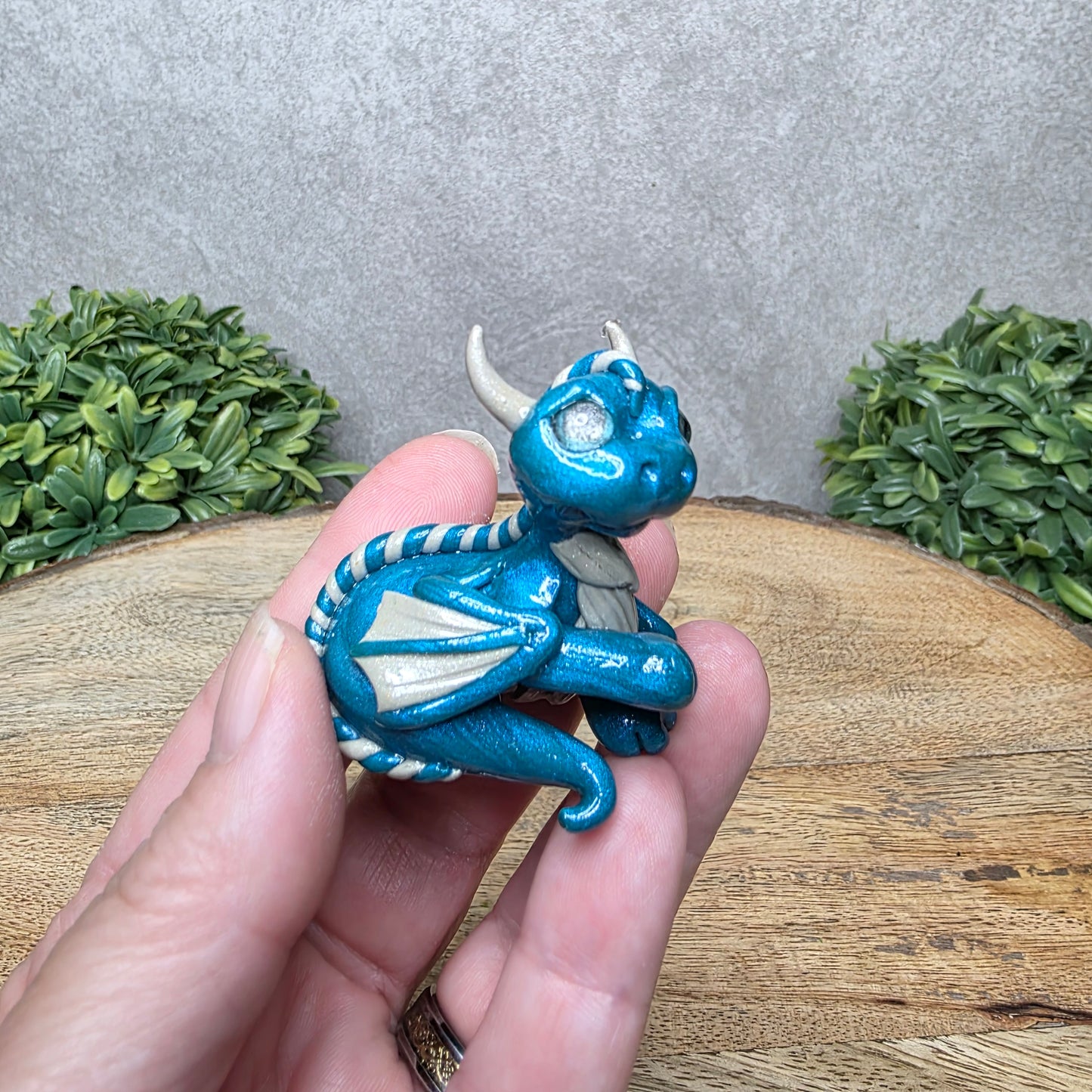A hand holding a baby clay dragon against a wood background with greenery