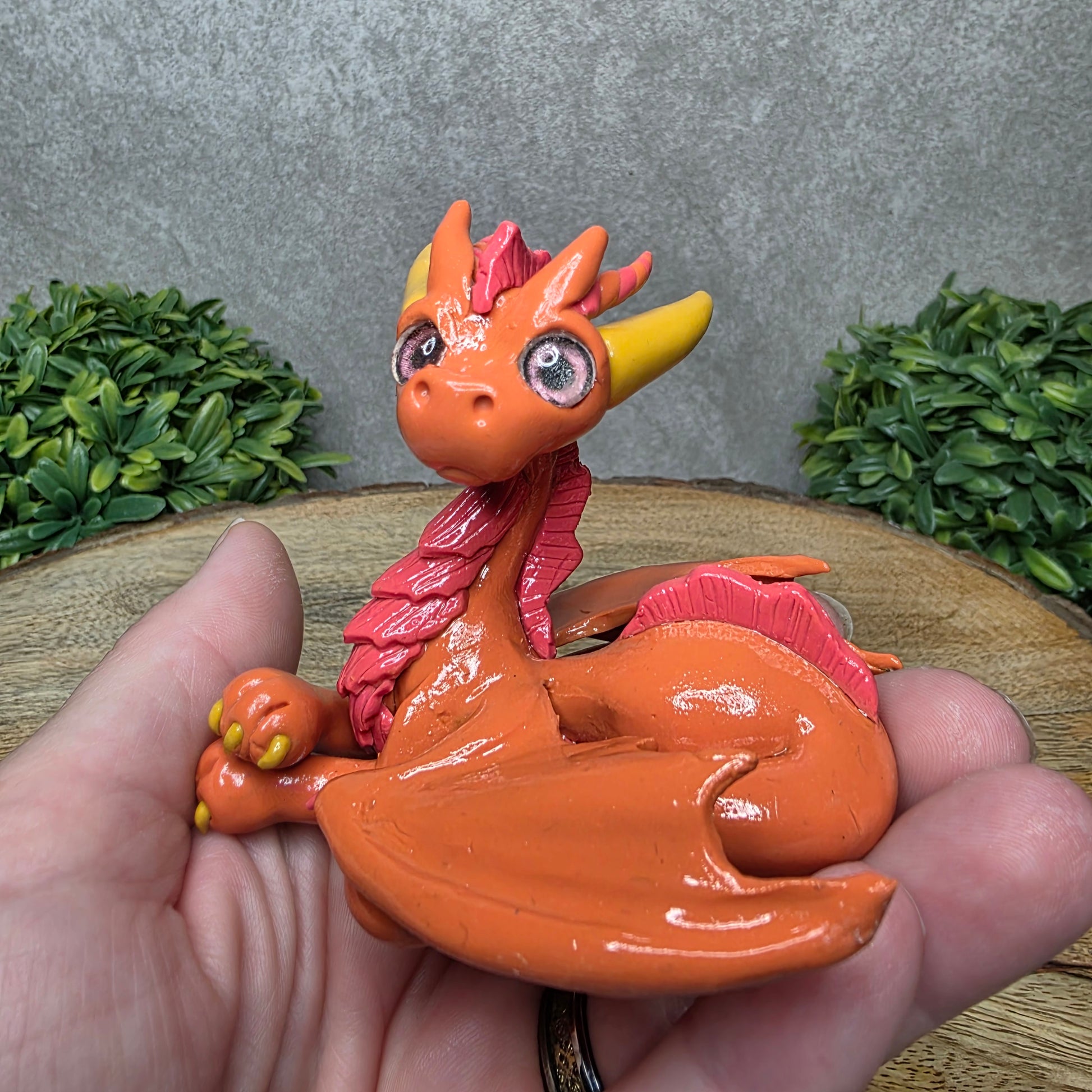 A hand holding a clay dragon against a wood background with greenery