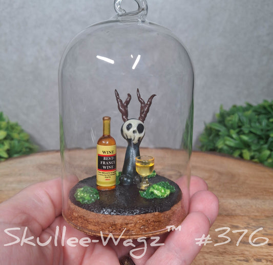 Glass dome with a skull faced creature with moss and wine bottle, held by a hand with a blurred green background. Text says Skullee-Wagz #376