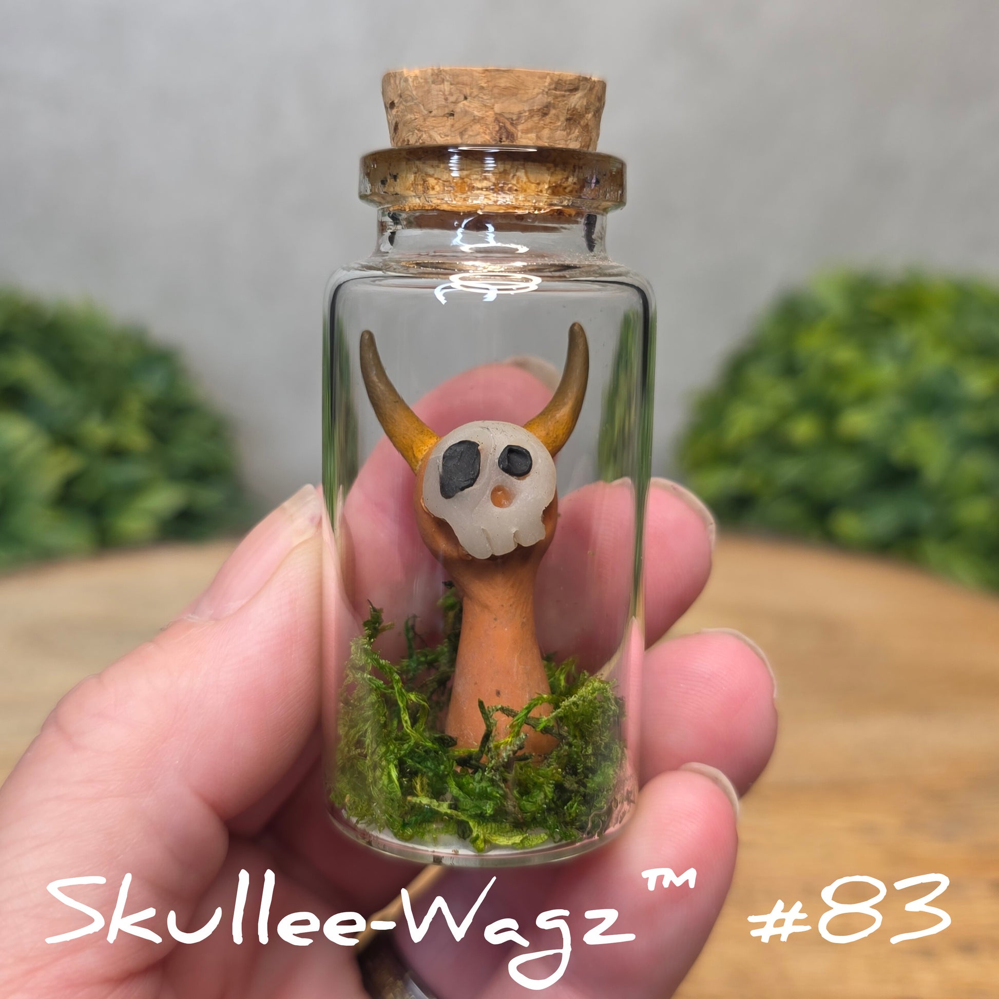 Hand holding a skull faced creature in a jar against a wood background with greenery