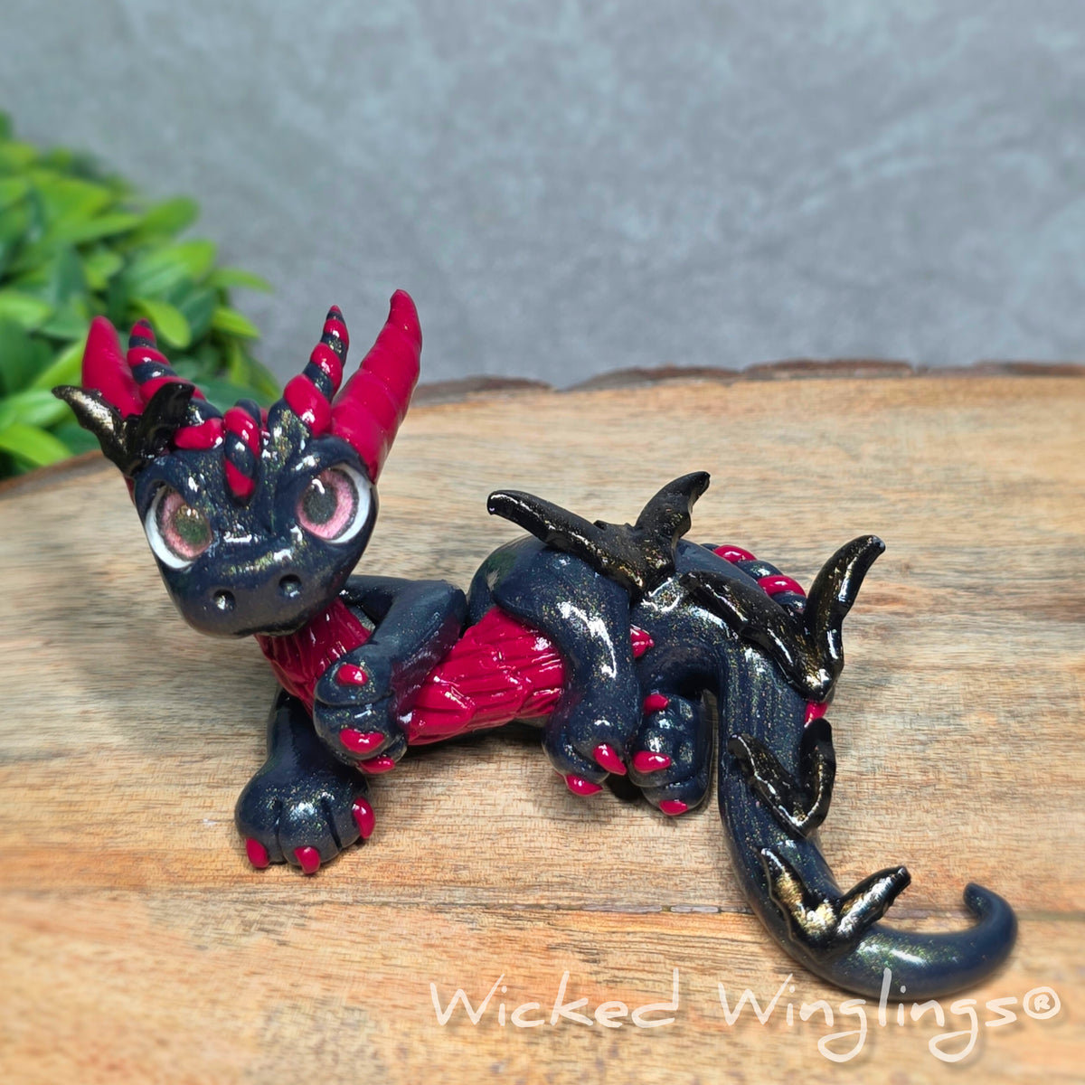 A clay dragon with bats sitting on a wood surface with greenery. Text reads Wicked Winglings.