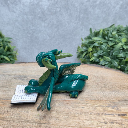 A dragon reading a book against a wood surface with greenery
