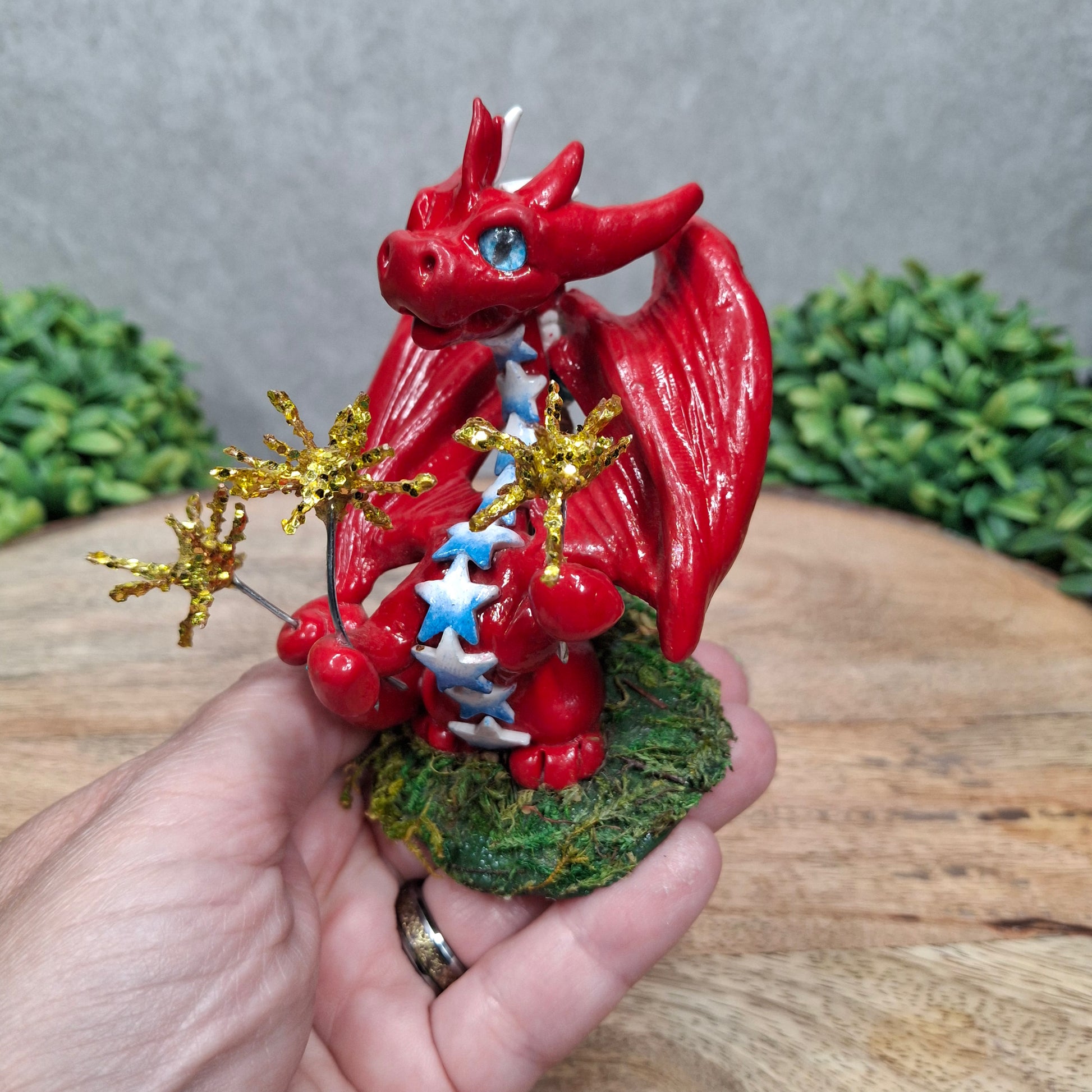A hand holding a clay dragon with sparklers against a wood surface with greenery