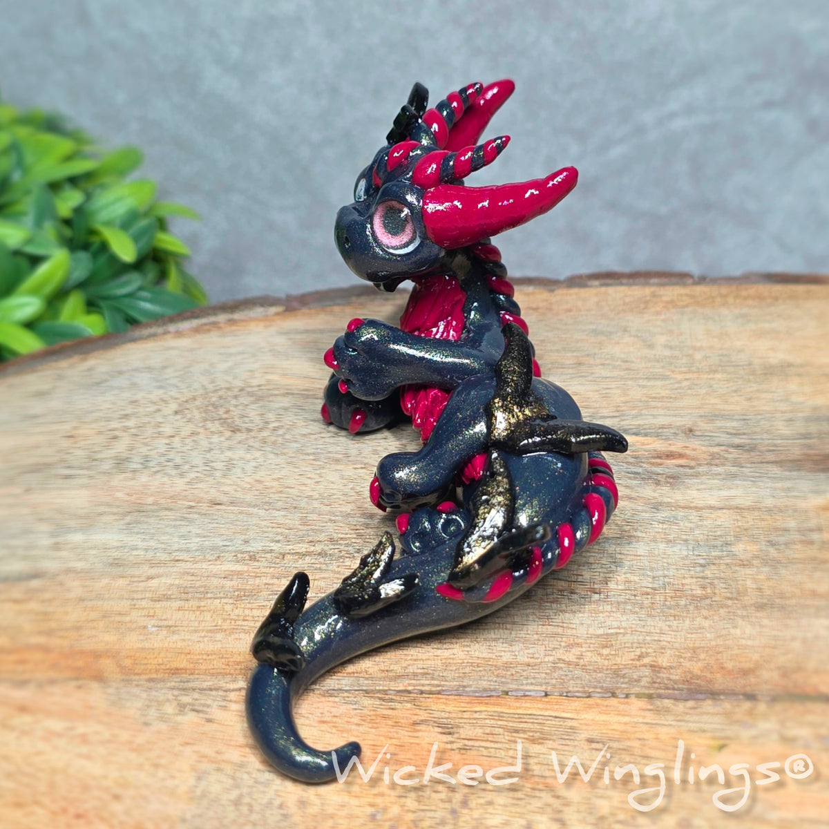 A side view of a clay dragon with bats sitting on a wood surface with greenery. Text reads Wicked Winglings.