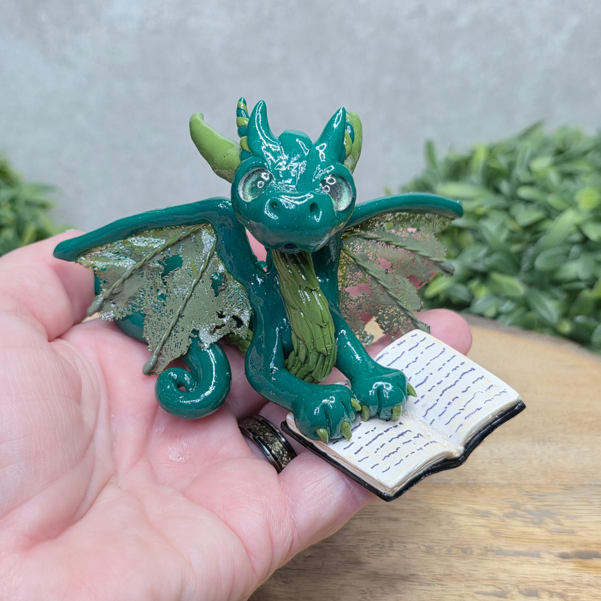 Hand holding a dragon reading a book against a wood surface with greenery