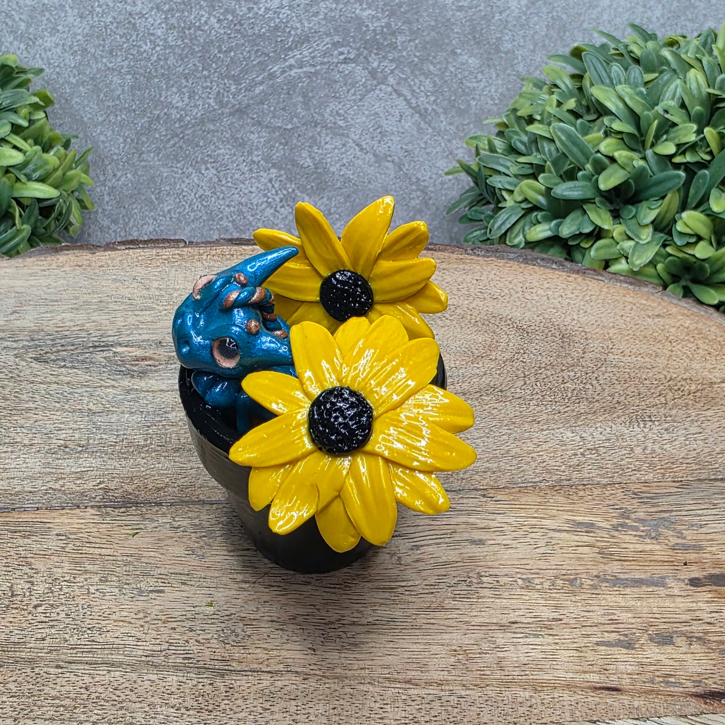 A side view of a clay baby dragon in a pot with black eye susans against a wood surface with greenery.