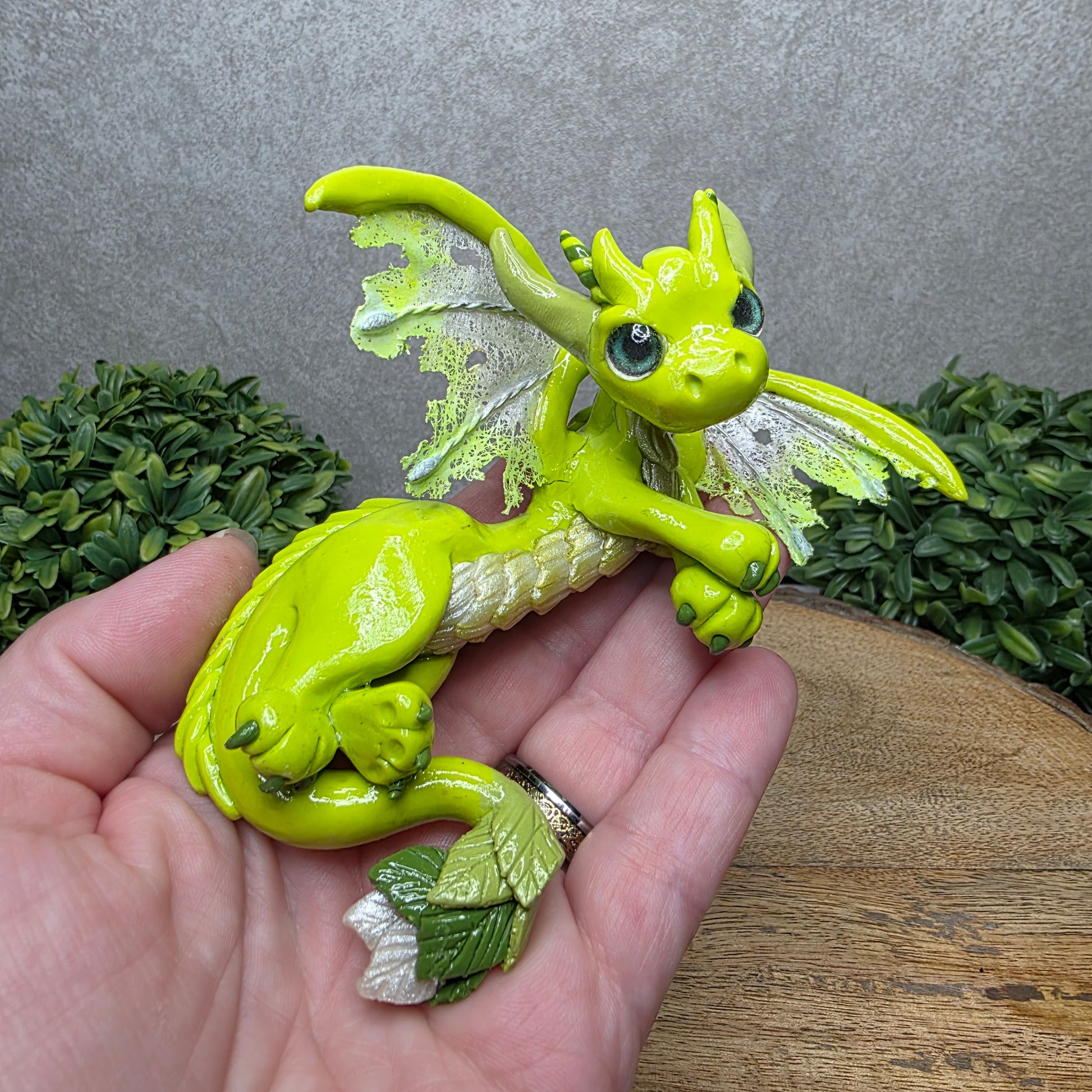 A hand holding a clay dragon against a wood background with greenery