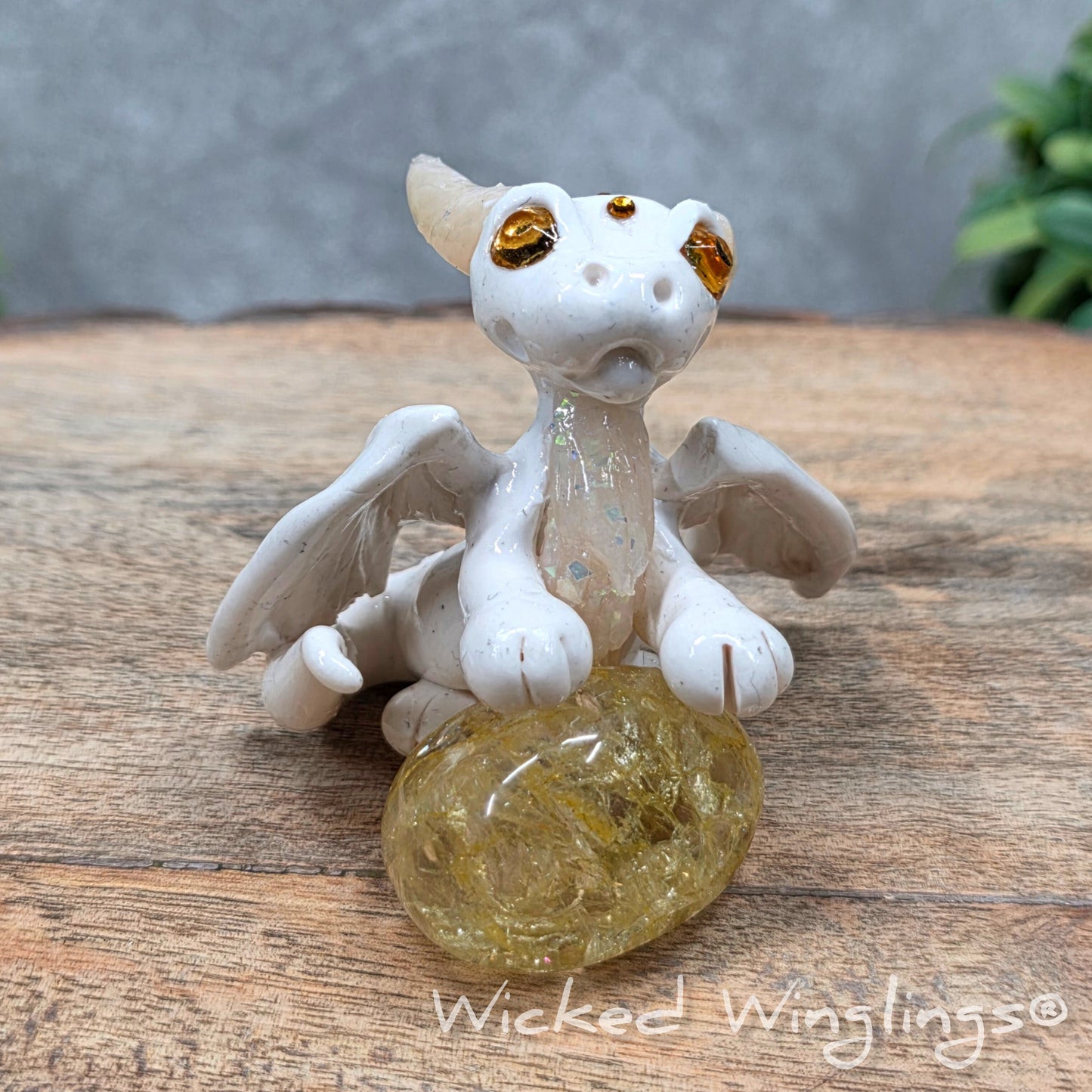 A baby clay dragon sculpture with a crackled quartz against a wood background with greenery and text Wicked Winglings