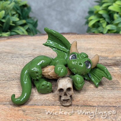 A clay dragon with a skull on a wood surface with greenery. Includes text Wicked Winglings