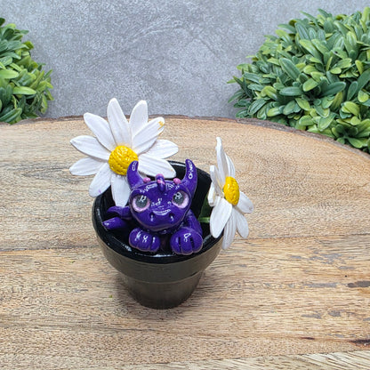 A clay baby dragon in a pot with daisies against a wood surface with greenery.