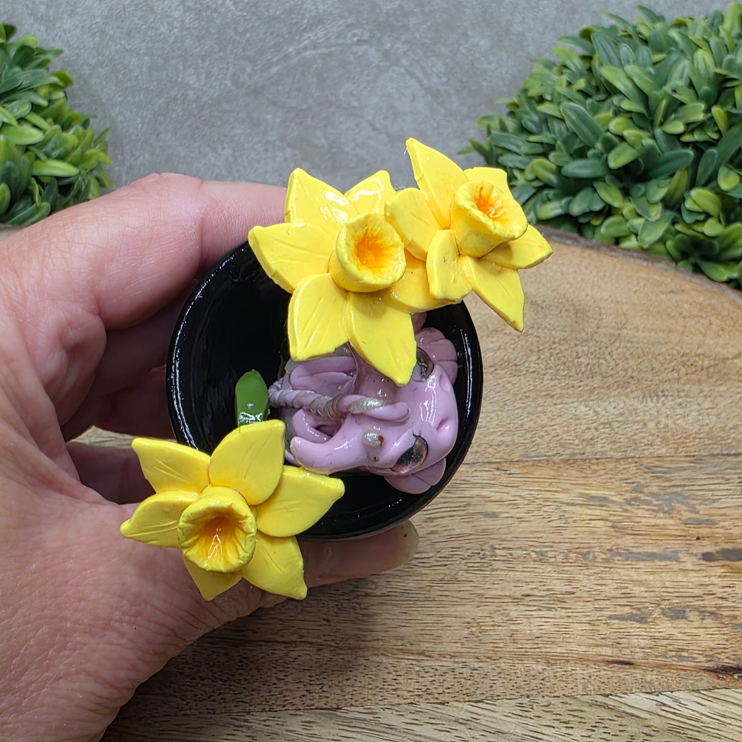Top view of a hand holding a clay baby dragon in a pot with daffodils against a wood surface with greenery.
