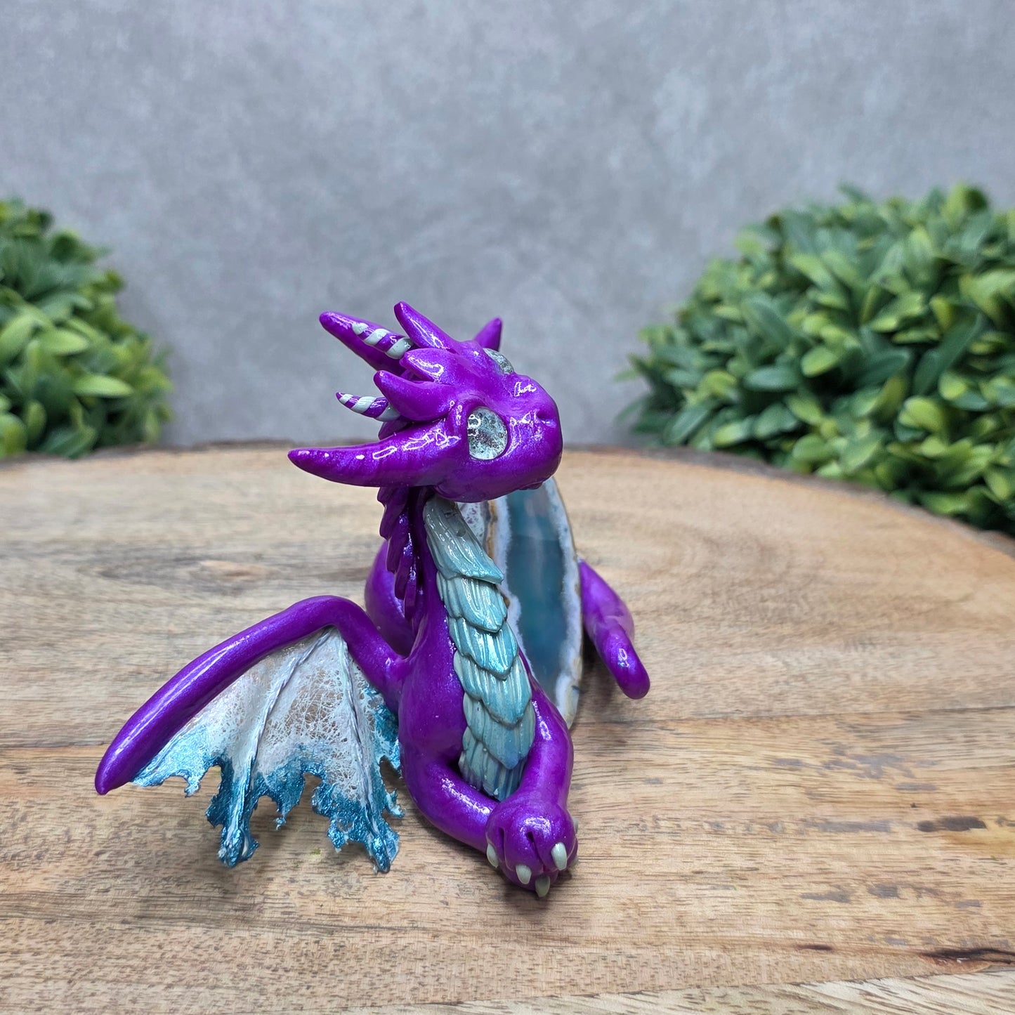 Side view of a clay dragon with an agate slice against a wood surface with greenery.