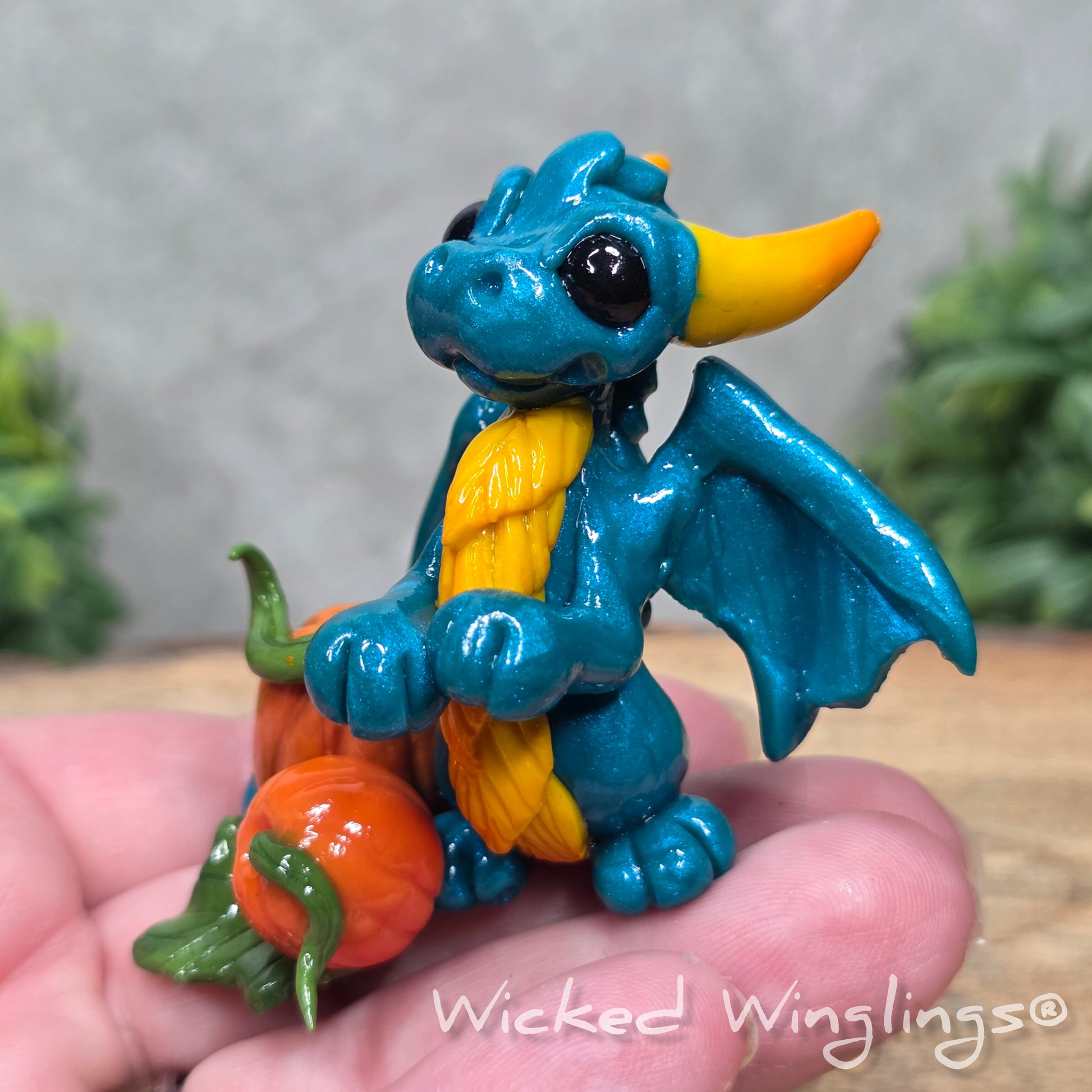 A hand holding a baby clay dragon sculpture with pumpkins against a wood background with greenery and text Wicked Winglings