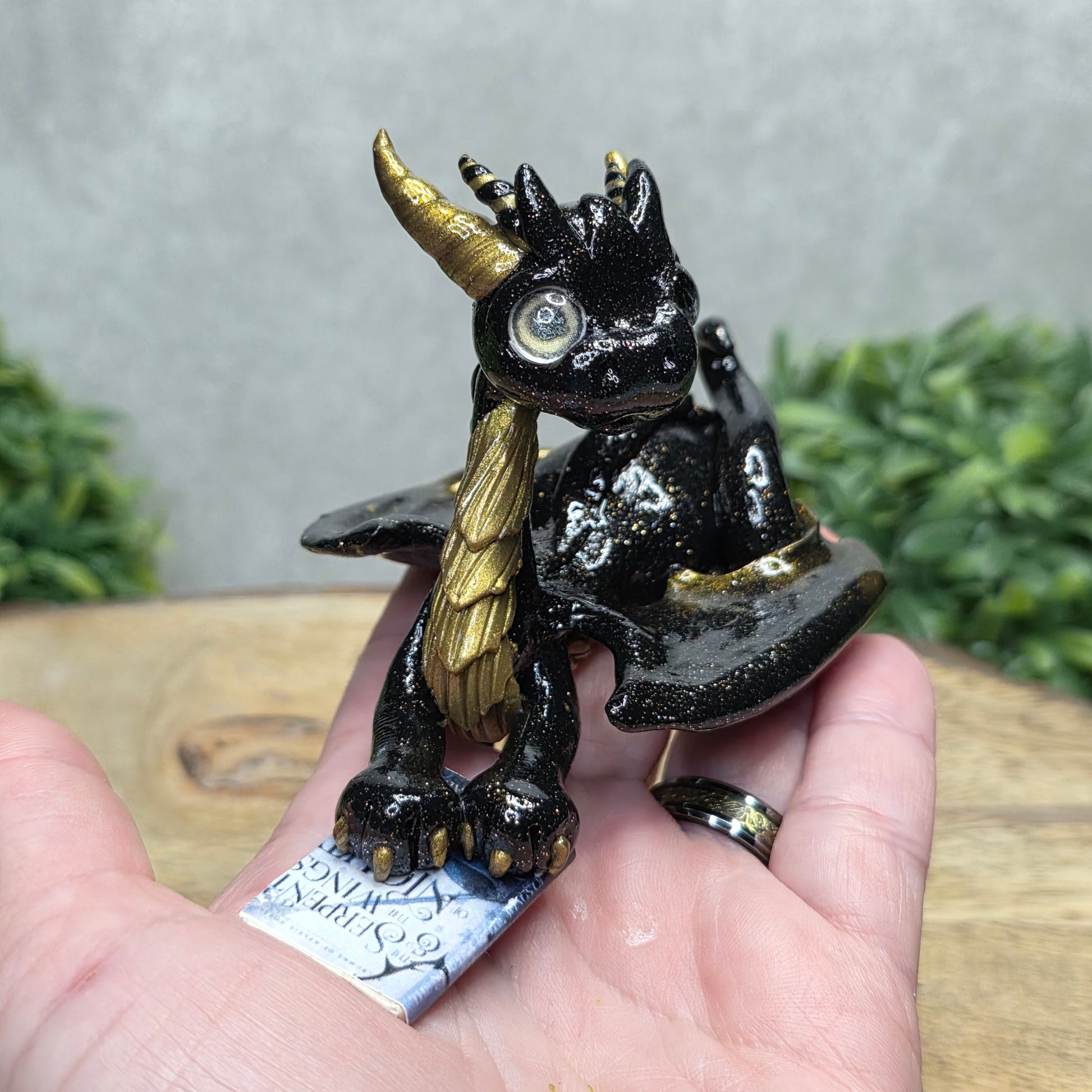 Hand holding a dragon reading a book against a wood surface with greenery