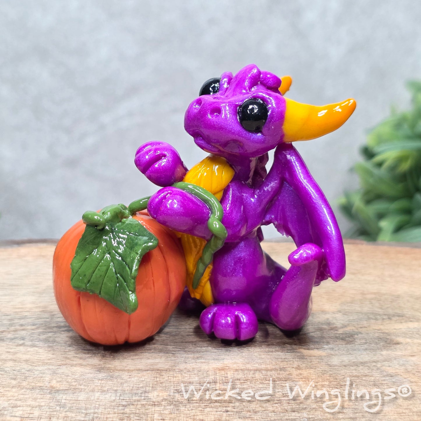 A baby clay dragon sculpture with a pumpkin against a wood background with greenery and text Wicked Winglings