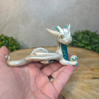 A hand holding a white and teal clay dragon against a blurred wood surface