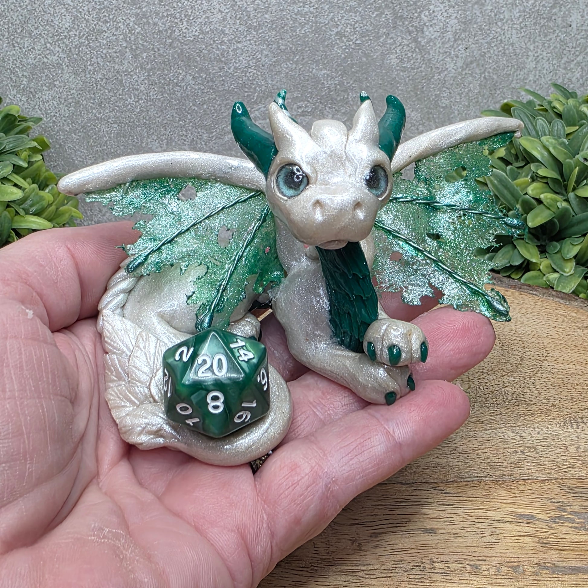 Hand holding a clay dragon with a D20 dice against a wood surface with greenery.