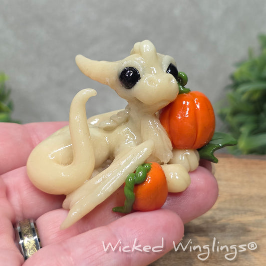 A hand holding a baby clay dragon sculpture with pumpkins against a wood background with greenery and text Wicked Winglings